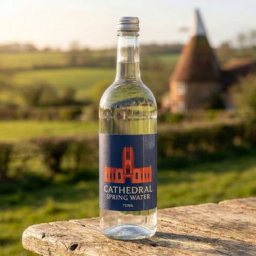 Cathedral Spring Water 750ml clear glass bottle with silver screw cap, navy label and red-orange Liverpool Cathedral silhouette, on an oak table in the Kent countryside